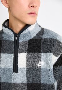 A Nike kockás mintás boleró fekete, szürke és fehér árnyalatokban, zippes nyakkal és hímzett logóval. Textúrált anyag.