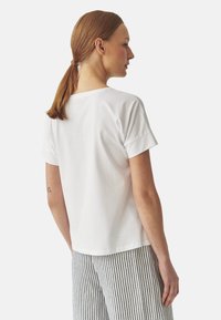 T-shirt en coton blanc avec une coupe légèrement ample, manches courtes retroussées et col rond, présenté de côté et de dos.