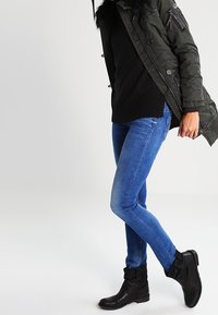 Olivinvihreä parka tekoturkistrimmin kanssa, päällä musta neule ja siniset kapeat farkut, yhdistettynä mustiin nilkkureihin.