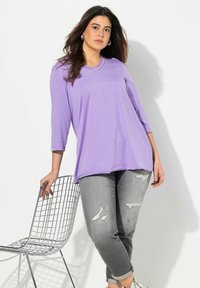 Lila långärmad topp med V-ringning, ihop med grå slitna jeans. Modellen lutar sig mot en metallstol och visar upp den avslappnade passformen.