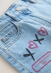 Poche de jean en denim bleu clair avec un X noir et un cœur rose ainsi qu'un texte tamponné rouge près de la couture et une fermeture par bouton métallique.
