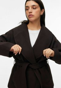 Abrigo marrón oscuro con cinturón y solapa ancha, hecho de un material suave. Presenta mangas largas y un corte holgado con bolsillos laterales.