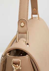 Sac à main en cuir beige avec garnitures dorées, fermeture éclair visible et logo LJ embossé sur la fixation de la sangle.