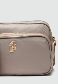 Geantă din nailon bej cu o textură netedă, având un fermoar auriu și un accent cu logo embosat. Design compact cu mai multe compartimente cu fermoar.