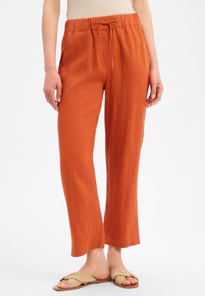 Vrouw draagt losse, verbrande oranje trekkoordbroek met zijzakken en beige platte sandalen, staand tegen een witte achtergrond.