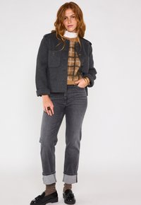 Veste en laine grise avec deux poches avant, superposée à un pull à carreaux marron. Assortie avec un jean gris et des mocassins noirs, mettant en valeur des poignets retroussés.
