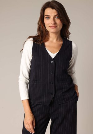 Marineblauw streeppatroon vest met vijf knopen, met een geknipt kraag en twee voorzakken, gedragen over een wit lang mouwtop. Gladde textuur.