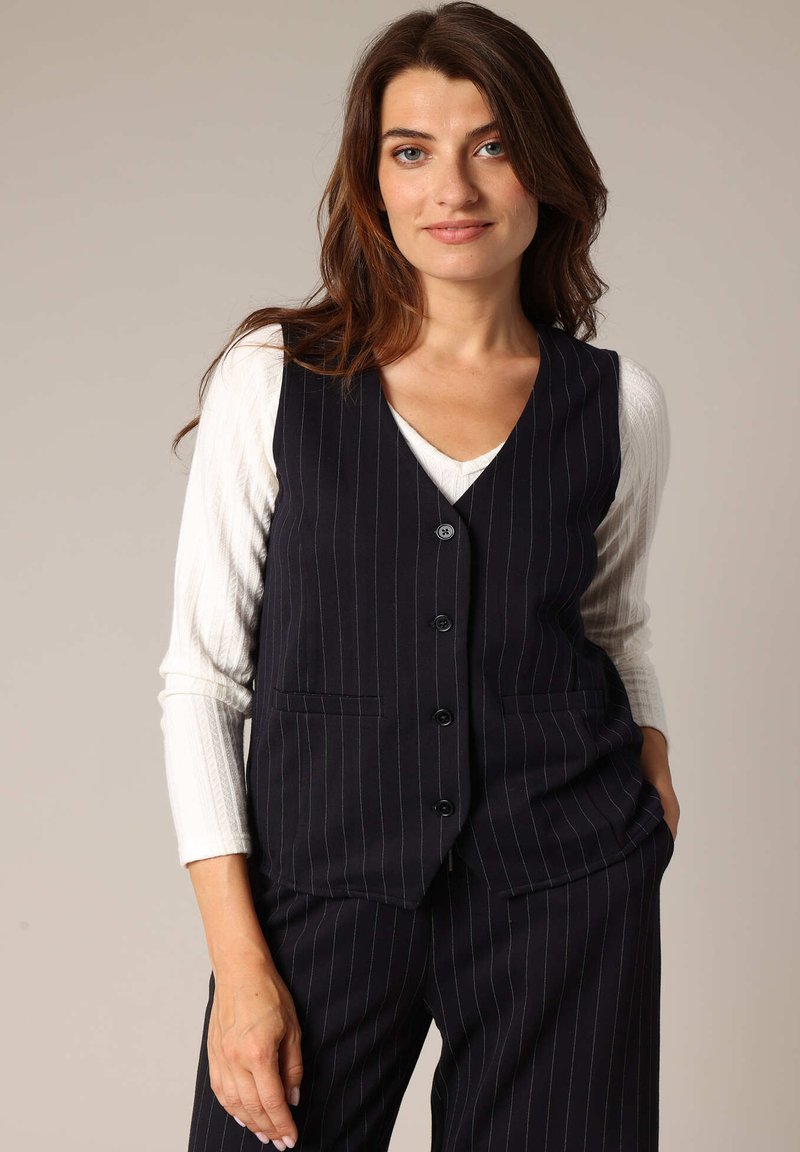 Marineblauw streeppatroon vest met vijf knopen, met een geknipt kraag en twee voorzakken, gedragen over een wit lang mouwtop. Gladde textuur.