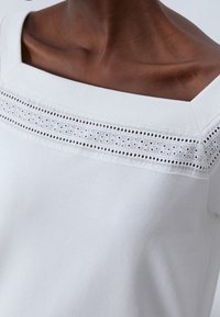 Vit topp med fyrkantig halsringning och dekorativ öljettkant över bröstet. Slät tyg med en mjuk textur.