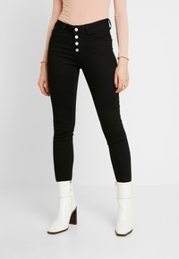 Jeans skinny negros con una cremallera de cinco botones plateados, bolsillos frontales y una textura suave. Combinados con botas blancas hasta el tobillo y una blusa rosa de canalé.