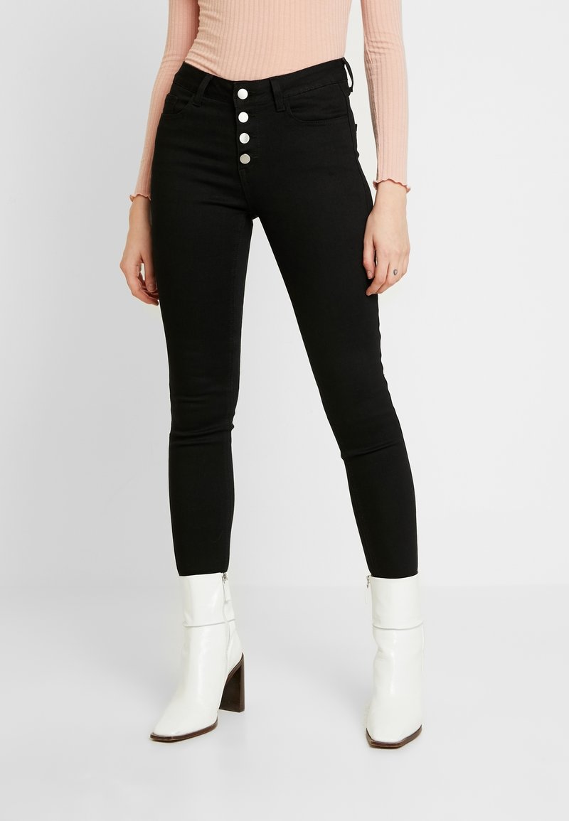 Jeans skinny negros con una cremallera de cinco botones plateados, bolsillos frontales y una textura suave. Combinados con botas blancas hasta el tobillo y una blusa rosa de canalé.