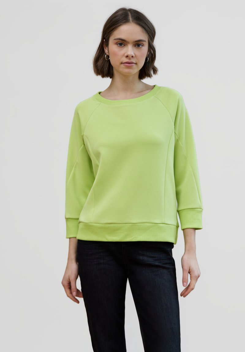 Sudadera verde clara con mangas raglán tres cuartos, puños y dobladillo acanalados. Textura suave y ajuste informal, combinada con vaqueros oscuros.