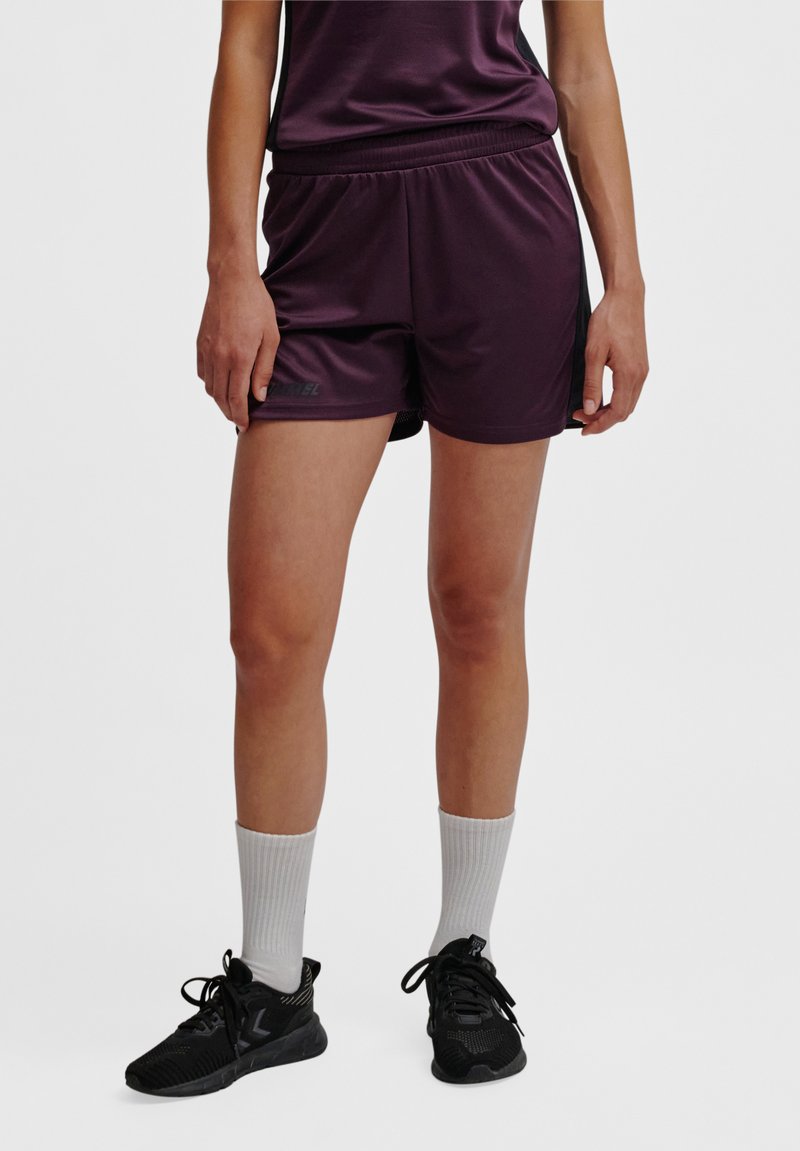 Person trägt dunkelviolette Sportshorts, passendes Oberteil, weiße Kniestrümpfe und schwarze Laufschuhe, steht vor einem einfarbigen Hintergrund.