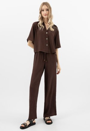 Młoda kobieta z długimi blond włosami, ubrana w luźną brązową koszulę zapinaną na guziki oraz pasujące szerokie spodnie, nosi czarne sandały w panterkę.