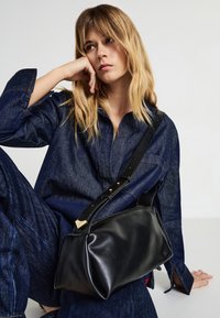 Borsa in pelle nera con forma strutturata, dotata di un manico corto e di una tracolla lunga regolabile, completata da accenti in metallo dorato.