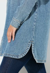 Lichtblauw denimjack met een ronde zoom, contrasterende stiksels en een knoopsluiting bij de mouwen. G gedragen over donkerblauwe spijkerbroek.