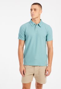 Polo shirt in licht teal stof met korte mouwen, geribbelde kraag en een knoopsluiting met drie knopen. Draagt met beige gestructureerde shorts.