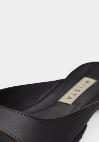 Μαύρο δερμάτινο παπούτσι τύπου slip-on με χαμηλό τακούνι και ορατή ετικέτα στο εσωτερικό που γράφει "MISTAA" σε λευκό φόντο.