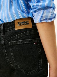 Mustmustestega teksad on mustad ja neil on pruun nahklabel, millel on kiri "TOMMY HILFIGER." Osaliselt on nähtav helesinine triibuline särk.