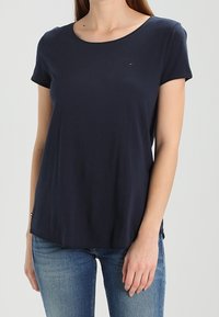 Žena v jednoduchém tmavě modrém tričku s krátkým rukávem a modrých džínách, stojící na světle šedém pozadí.