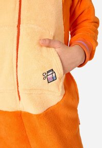 Oranžno in smetanasto enodelno oblačilo iz flisa z zadrgo spredaj, kenguru žepom in vezenjem mlečne embalaže. Mehka tekstura in sproščen kroj.