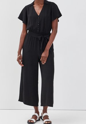 Femme portant une combinaison noire à manches courtes avec des boutons et une taille nouée, associée à des sandales blanches et un bracelet au poignet gauche.