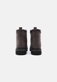 Un paio di stivali in pelle marrone scuro con suole spesse nere e linguette sul tallone, mostrati da dietro su uno sfondo bianco.