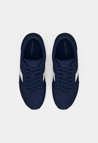 Σκούρα μπλε αθλητικά παπούτσια με λείο επάνω μέρος, λευκές πλαϊνές λεπτομέρειες, στρογγυλεμένη μύτη και σχέδιο με κορδόνια. Το λογότυπο είναι ορατό στο εσωτερικό πέλμα.