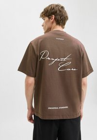 Brun bomulds t-shirt med korte ærmer, med hvid kursiv tekst "Project Care" på ryggen og "UNIVERSAL STANDARD" ved kanten.