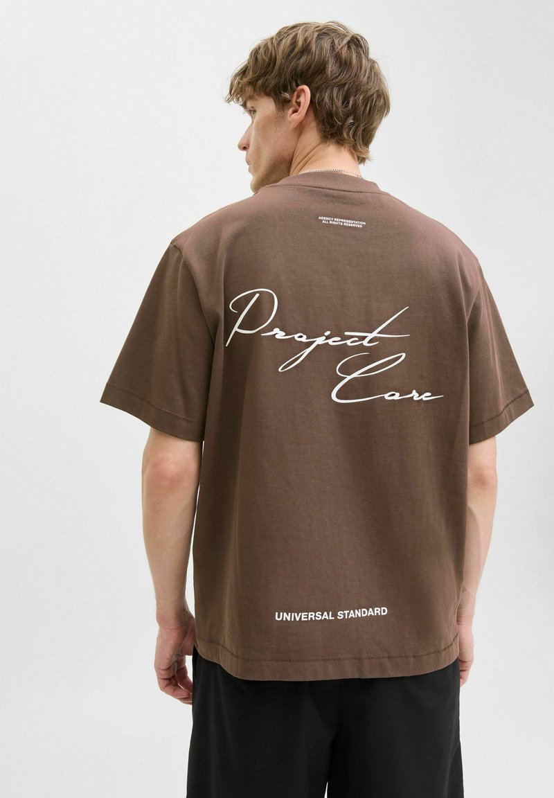 Brun bomulds t-shirt med korte ærmer, med hvid kursiv tekst "Project Care" på ryggen og "UNIVERSAL STANDARD" ved kanten.
