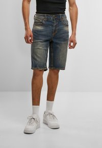 Muž v vyblednutých modrých džínsových šortkách, bielych ponožkách a bielych teniskách, stojaci pred jednoduchým svetlošedým pozadím.