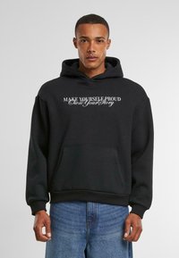 Sort hætte sweatshirt med forlommer og hvid tekst, der siger "GØR DIG SELV STOLTHED - Ejer din historie." Glat stof med ribbede mansjetter.