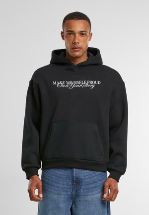 Sort hætte sweatshirt med forlommer og hvid tekst, der siger "GØR DIG SELV STOLTHED - Ejer din historie." Glat stof med ribbede mansjetter.