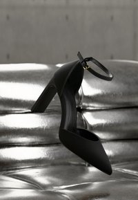 Μαύρο παπούτσι με ψηλό τακούνι και λουρί στο αστράγαλο, τοποθετημένο πάνω σε γυαλιστερό ασημί δερμάτινο ταπετσαρία με ραφή και καπιτονέ.