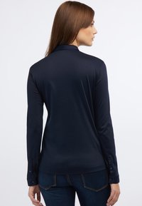 Navy long-sleeve shirt with a collar, smooth texture, tailored fit, and button details on the cuffs, paired with dark denim jeans.