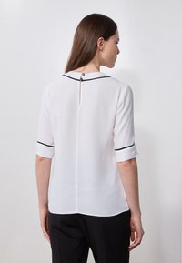 Blouse blanche à manches courtes, avec des accents en piped noirs le long du col et des manches, présentant un dos à trou de serrure avec une fermeture à bouton.