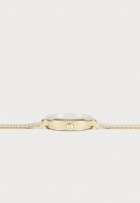 Reloj dorado con una caja redonda delgada, superficie lisa y una correa de malla metálica. Cuenta con un solo botón en el lateral para ajustar la hora.