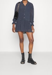 Kvinna i en marinblå prikkig minikjol, svarta ankelboots och bärande en svart axelväska med en guldkedjesnodd.