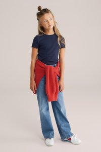 Námornicky modré žebrované tričko s krátkým rukávem a vlnitými okraji, spárované se světle modrými širokými džíny a červeným pleteným svetrem uvázaným kolem pasu. Bílé tenisky.