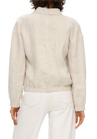 Osoba w beżowej koszuli z długim rękawem z elastycznymi mankietami i dołem, w połączeniu z białawymi spodniami o wysokim stanie, widziana z tyłu.