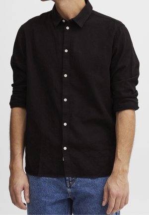 Homme portant une chemise noire à manches longues boutonnée avec les manches retroussées, associée à un jean en denim bleu, debout devant un fond uni.