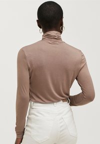 Langærmet turtleneck i lysebrunt stof med glat tekstur, parret med hvide højtaljede bukser med baglommer og etiketsyning.