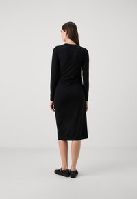 Vestido preto de manga longa com silhueta ajustada, comprimento midi, textura suave e detalhe franzido nas costas. Combinado com sapatos pretos.