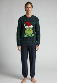 Zeleno-černý károvaný top s dlouhým rukávem, který zobrazuje vyšitý grafický motiv Grinche se červenou santovskou čepicí, doplněný kalhotami s elastickými manžetami v námořnické modři.