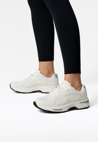 Białe buty sportowe z teksturowanej siateczki i syntetycznymi akcentami, z wygodną podeszwą i czarną gumową bieżnią. W połączeniu z czarnymi legginsami.