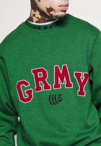 Felpa verde con lettering rosso "GRMY", caratterizzata da un colletto nero pattern e un dettaglio grafico ricamato sotto il testo.