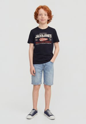Garçon roux debout, une main dans la poche, portant un t-shirt noir à motif graphique, un short en jean clair et des baskets bleu marine.