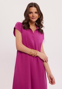 Fuksjowa sukienka z krótkimi rękawami i kołnierzykiem; miękka tekstura; charakteryzuje się luźnym krojem; zestawiona ze złotymi bransoletkami.