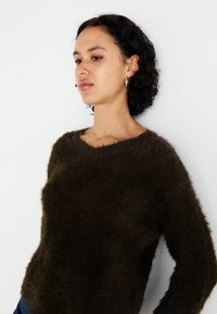 Dunkelgrüner, flauschiger Pullover mit Bootsausschnitt und langen Ärmeln. Verfügt über eine weiche Textur und eine lässige Passform. Das Modell posiert vor einem schlichten Hintergrund.