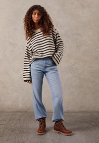 Pull oversized crème et noir rayé, jeans taille haute bleu clair et bottes marron à lacets avec une semelle robuste. Tenue texturée et décontractée.
